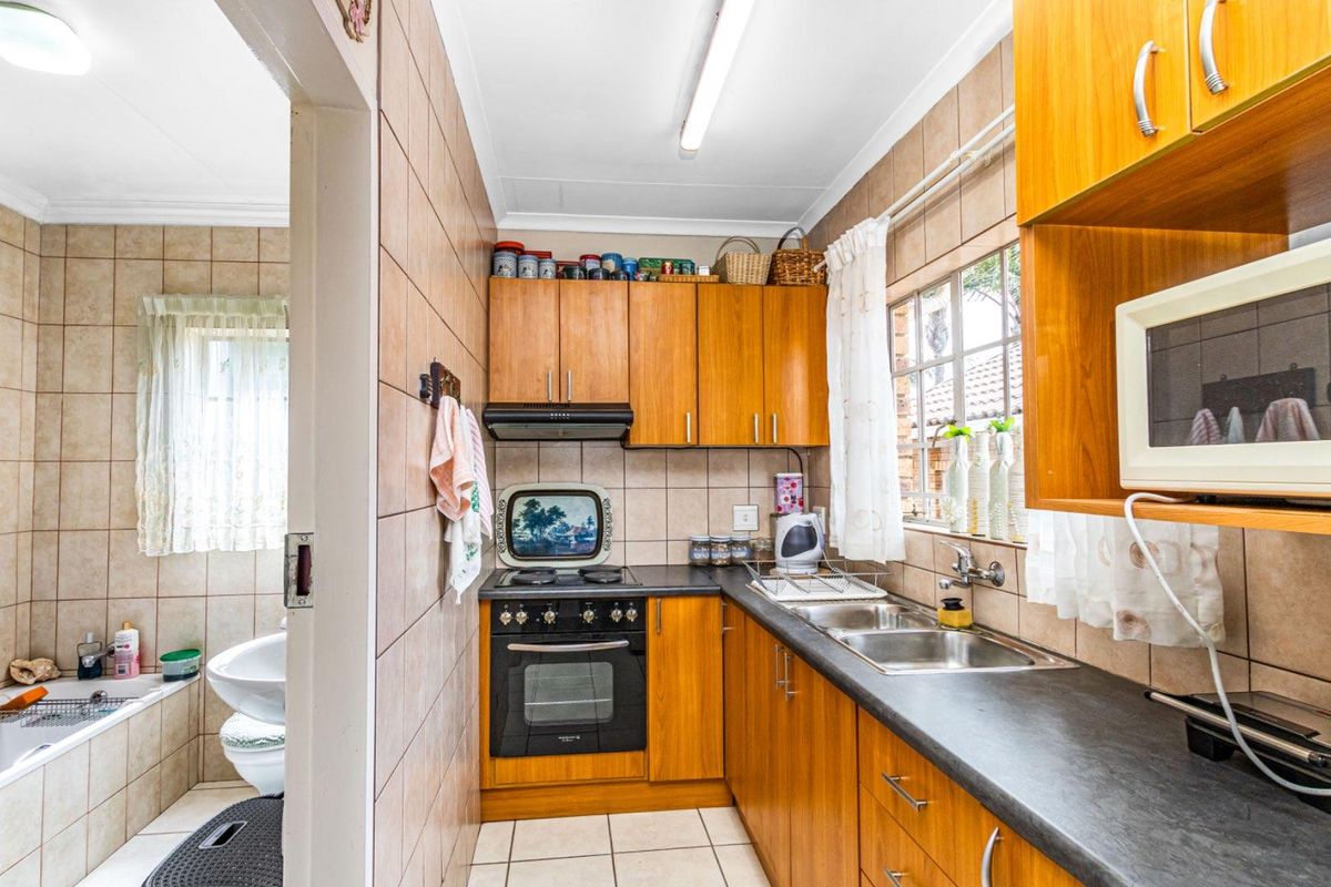 A compact kitchen featuring wooden cabinetry, a built-in oven and hob, and a double sink, with an adjacent bathroom visible through the doorway.