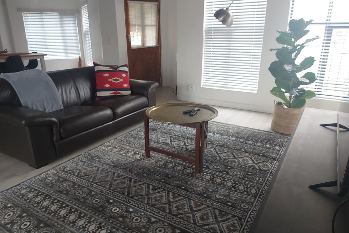 A bright and open living room featuring neutral laminate flooring, large windows with venetian blinds, and direct access to an exterior timber door.