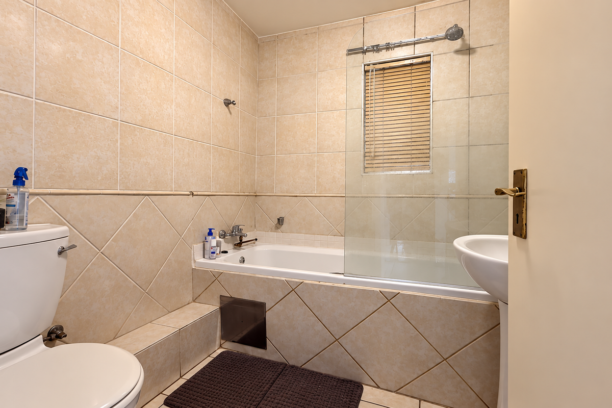 A functional bathroom featuring a bathtub with a glass shower screen, pedestal sink, toilet, and neutral ceramic wall and floor tiling.
