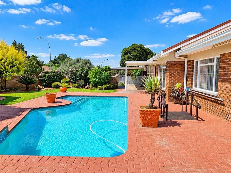 A well-maintained outdoor pool area featuring brick paving, a swimming pool, and a thatched gazebo, set against a brick-clad house exterior.