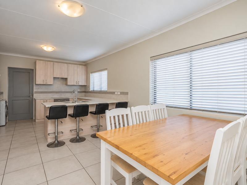 Open-plan kitchen and dining area featuring light wood cabinetry, a breakfast bar island, tiled flooring, and large windows with venetian blinds.