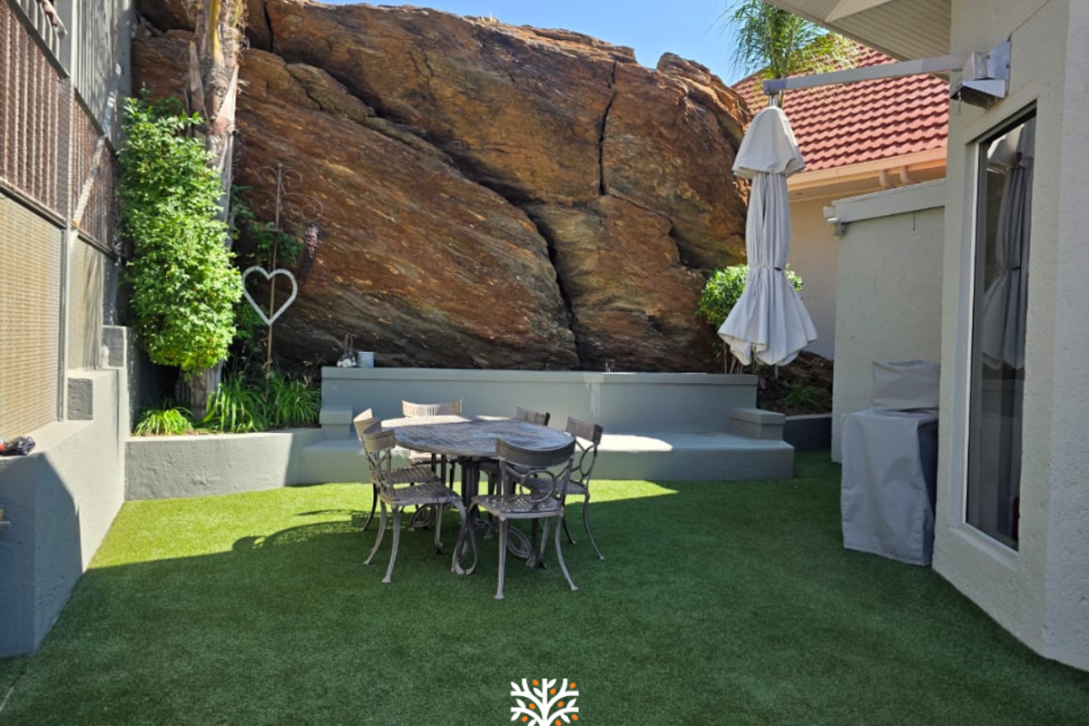 A private rear courtyard featuring artificial turf, a built-in retaining wall, and a unique natural rock face backdrop.