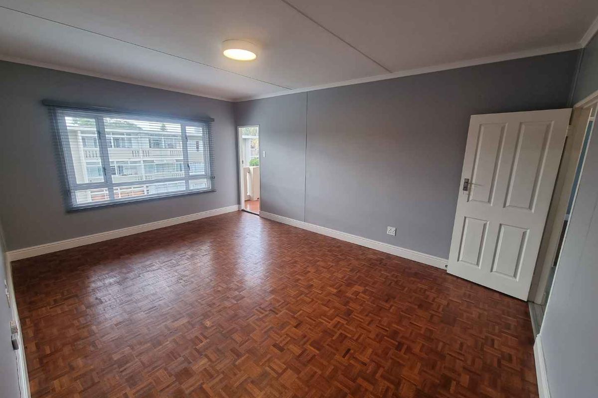 A spacious, empty bedroom featuring classic parquet flooring, neutral grey walls, and a large window providing natural light.