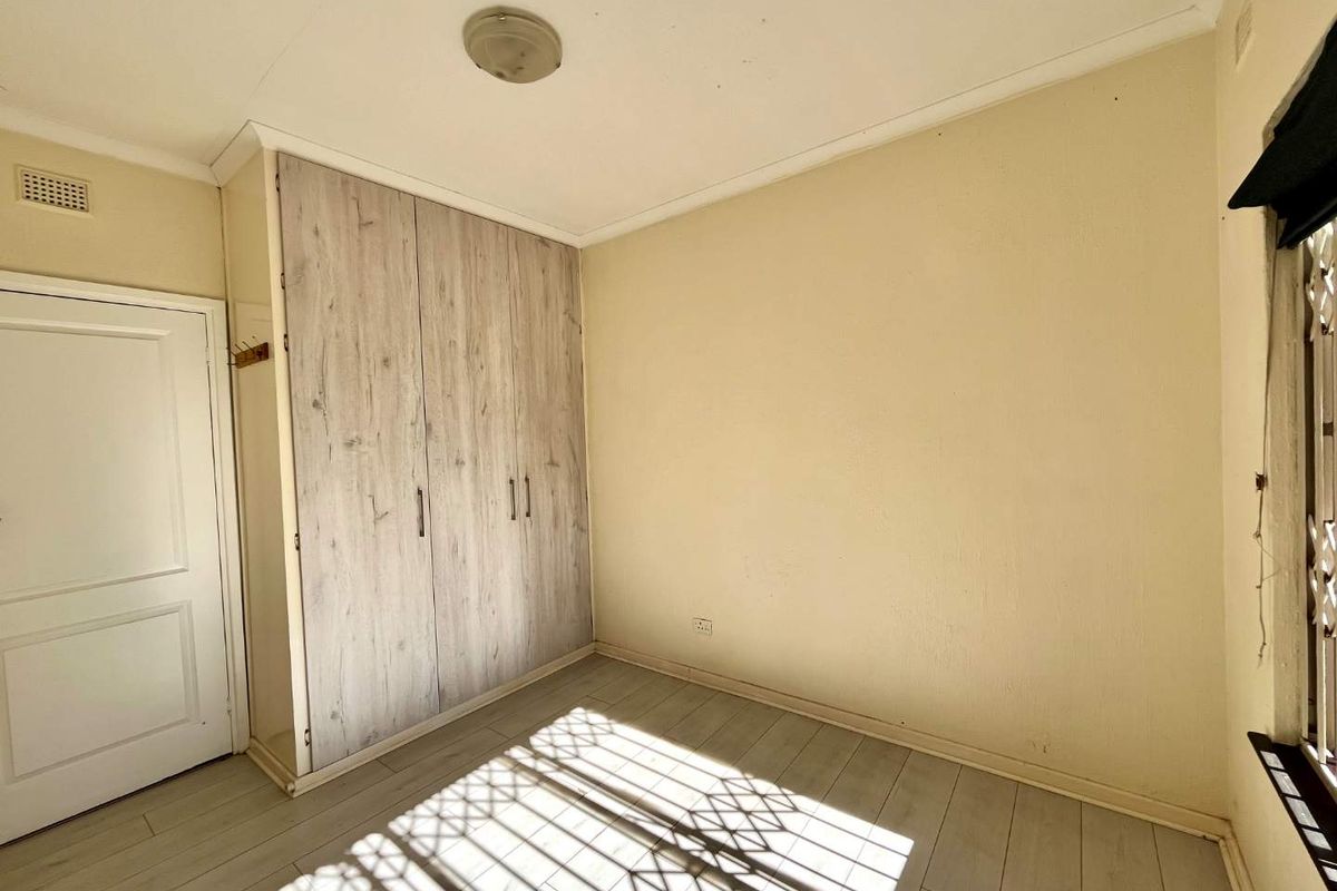 A vacant bedroom featuring light-toned laminate flooring, built-in wardrobes with wood-grain finish, and a security gate on the window.