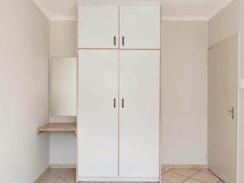 A clean, empty bedroom featuring a built-in wardrobe with overhead storage and an integrated vanity area, finished with neutral walls and tiled flooring.