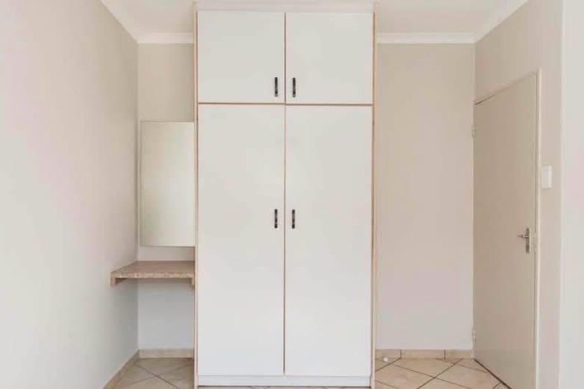 A clean, empty bedroom featuring a built-in wardrobe with overhead storage and an integrated vanity area, finished with neutral walls and tiled flooring.