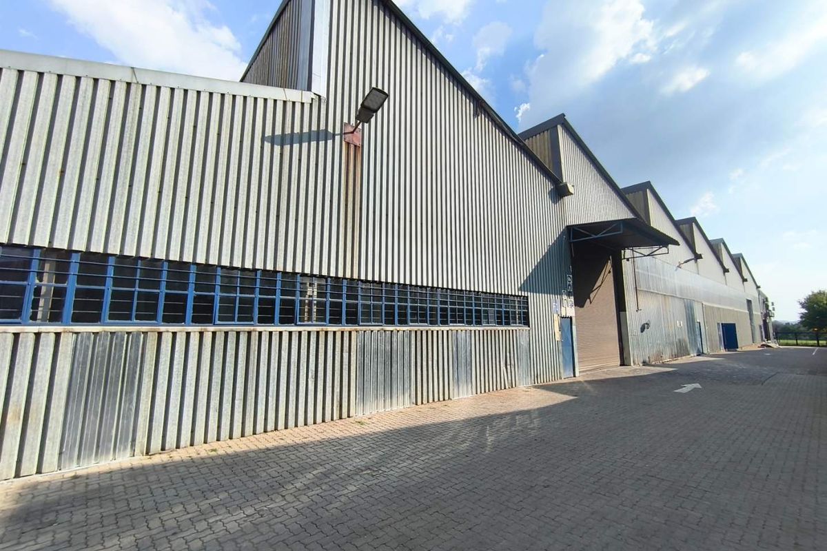 Industrial warehouse exterior featuring corrugated metal cladding, steel-framed windows, and a paved driveway with access via a large roller shutter door.