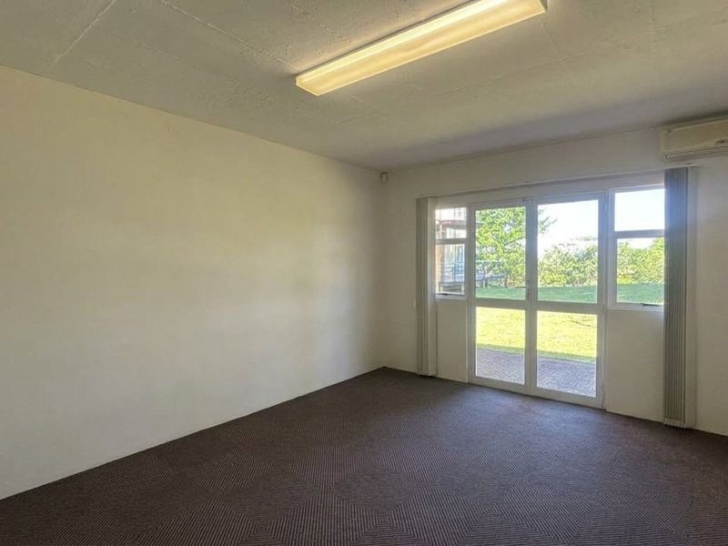 A vacant office space featuring carpet tiles, a split-system air conditioner, and a glass door providing natural light and garden views.