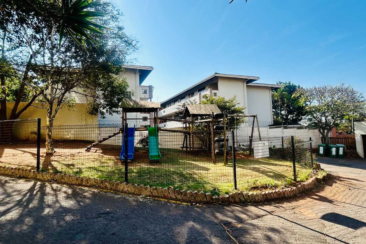 Communal outdoor area featuring a playground set, enclosed by security fencing and a stone retaining wall, situated between apartment blocks.