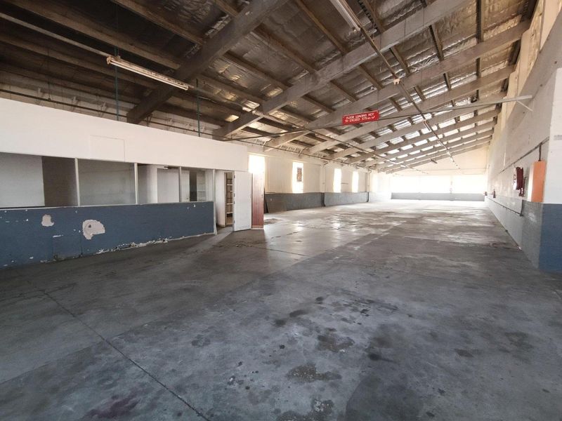 Spacious industrial warehouse area featuring exposed steel roof trusses, concrete flooring, and a partitioned office section, suitable for various commercial or storage uses.