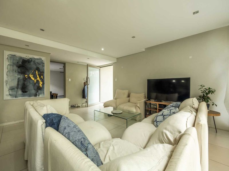 A spacious living room featuring neutral tiled flooring, recessed downlighting, and a glass door leading to an adjacent area.