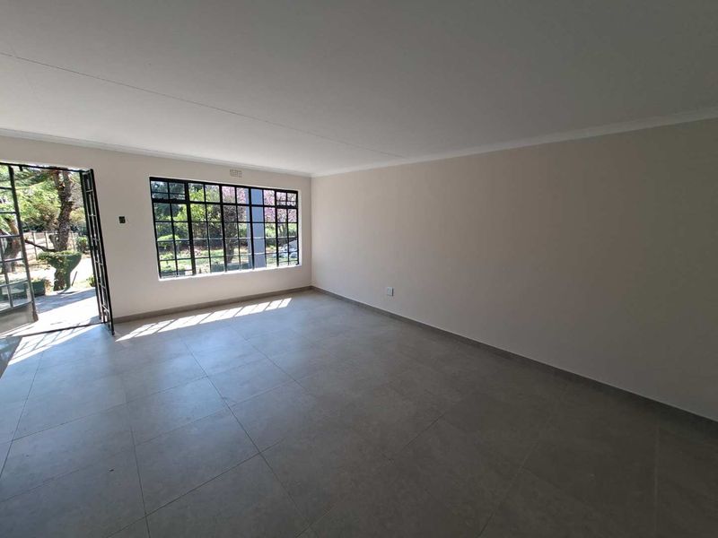 A bright, empty living room featuring neutral tiled flooring, coved ceilings, and large steel-framed windows with access to the exterior.