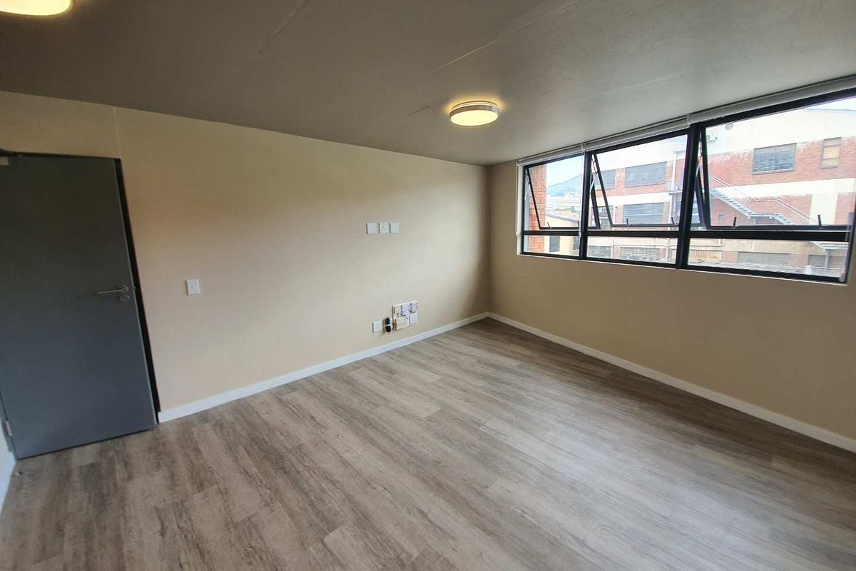 A bright, empty bedroom featuring modern laminate flooring, neutral wall tones, and large aluminium-framed windows providing natural light.