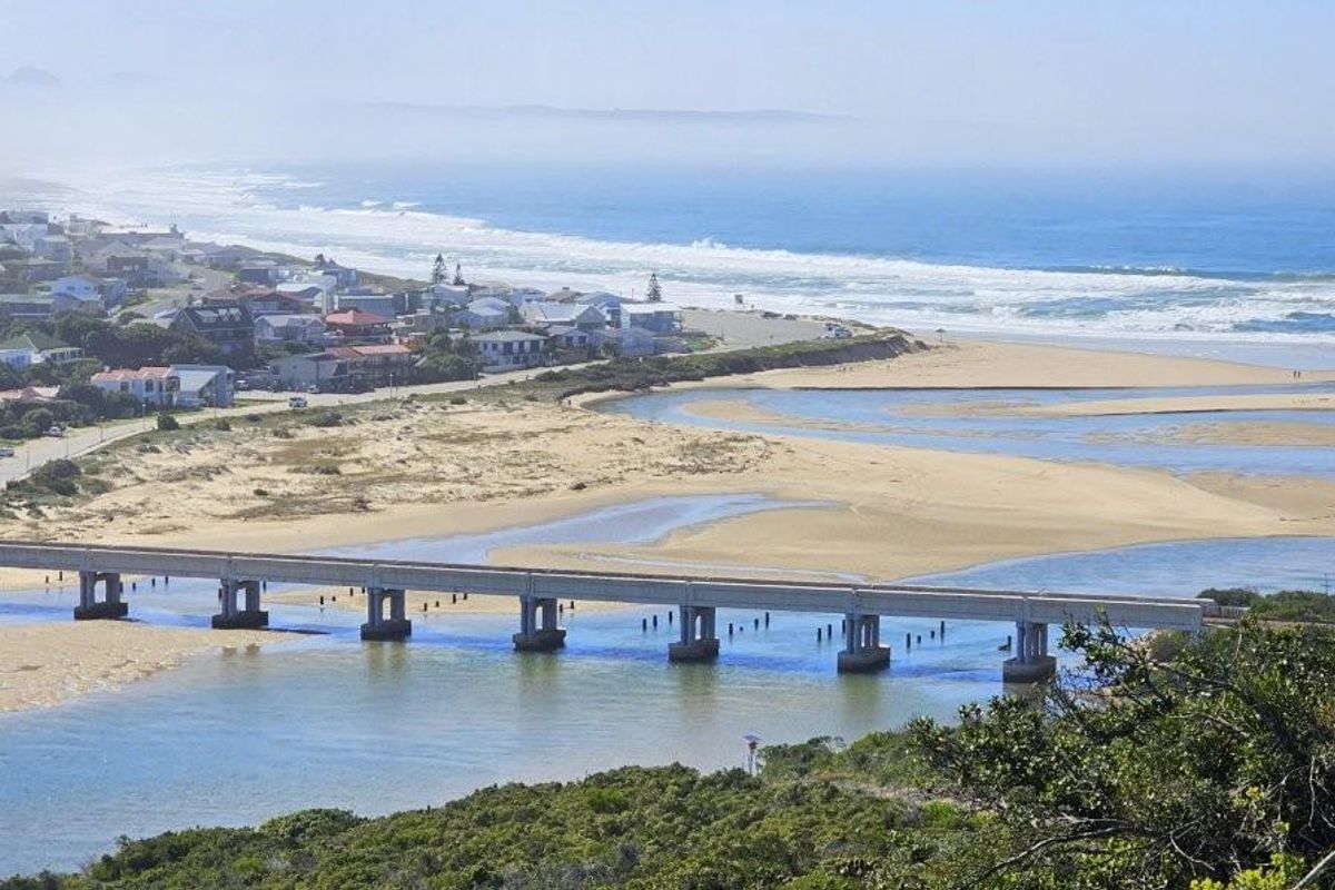 An elevated aerial view showcasing the coastal landscape, featuring a bridge spanning the river mouth, sandy beach, and surrounding residential area.