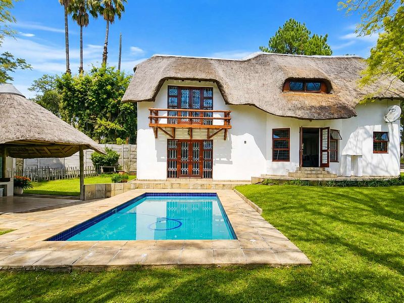 Rear view of a thatched-roof house featuring a swimming pool, a covered lapa with built-in braai, and a landscaped garden.