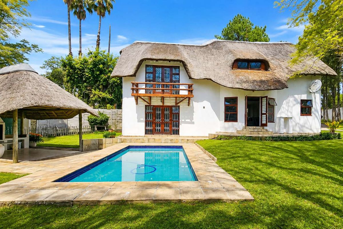 Rear view of a thatched-roof house featuring a swimming pool, a covered lapa with built-in braai, and a landscaped garden.
