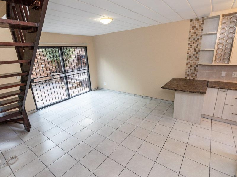 Open-plan living area featuring tiled floors, a wooden staircase, and a compact kitchen with laminate cabinetry and mosaic backsplash, leading to a private patio.