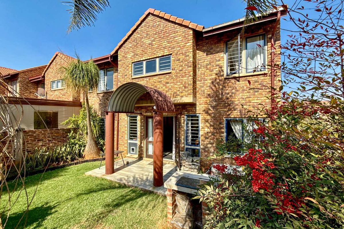 Rear view of a face brick townhouse featuring a paved patio, arched entrance canopy, and a built-in brick braai area set within a private garden.