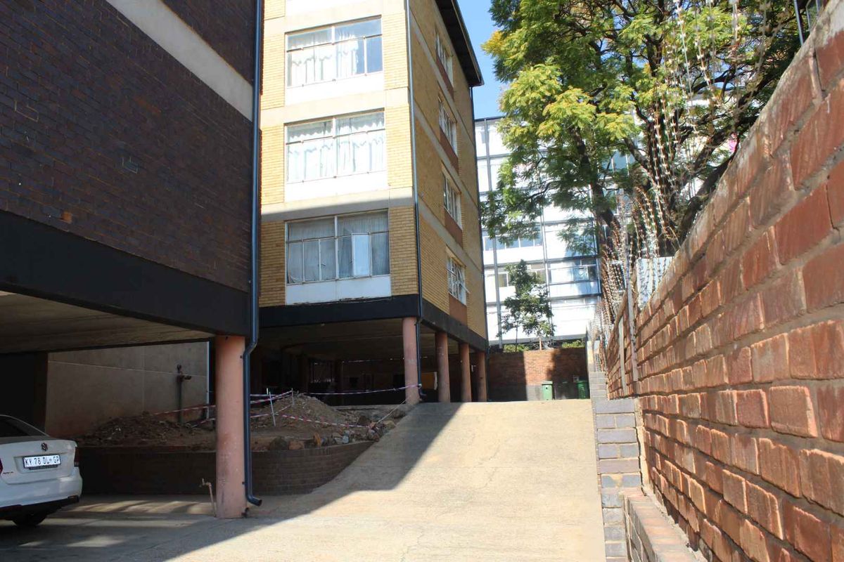Rear exterior view of the apartment building showing a concrete driveway, brick boundary wall, and ground-level stilt structure with ongoing site maintenance.