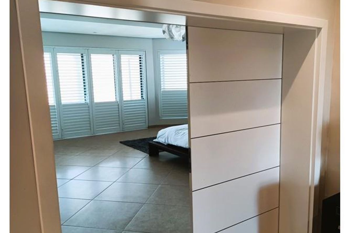 A view through a modern white sliding door into a spacious bedroom featuring large tiled flooring and plantation shutters.