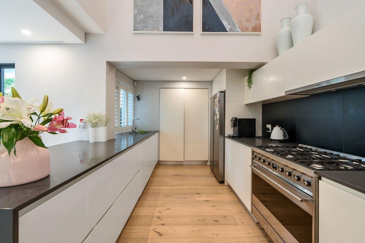 Modern galley-style kitchen featuring sleek white cabinetry, dark stone countertops, high-end gas range, and warm hardwood flooring.