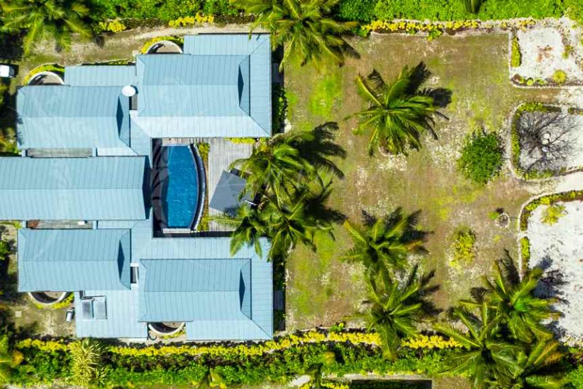 Aerial view of a villa property featuring a metal roof, central swimming pool, and landscaped garden grounds leading towards the beach.
