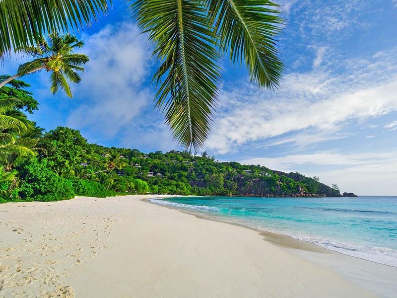 A scenic view of a pristine white sand beach with turquoise waters, lush tropical vegetation, and palm trees along the coastline.