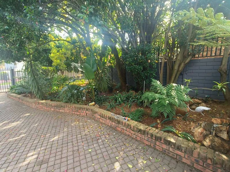 A landscaped garden area featuring a brick retaining wall, mature trees, and a paved driveway, enclosed by a painted masonry boundary wall and security fencing.