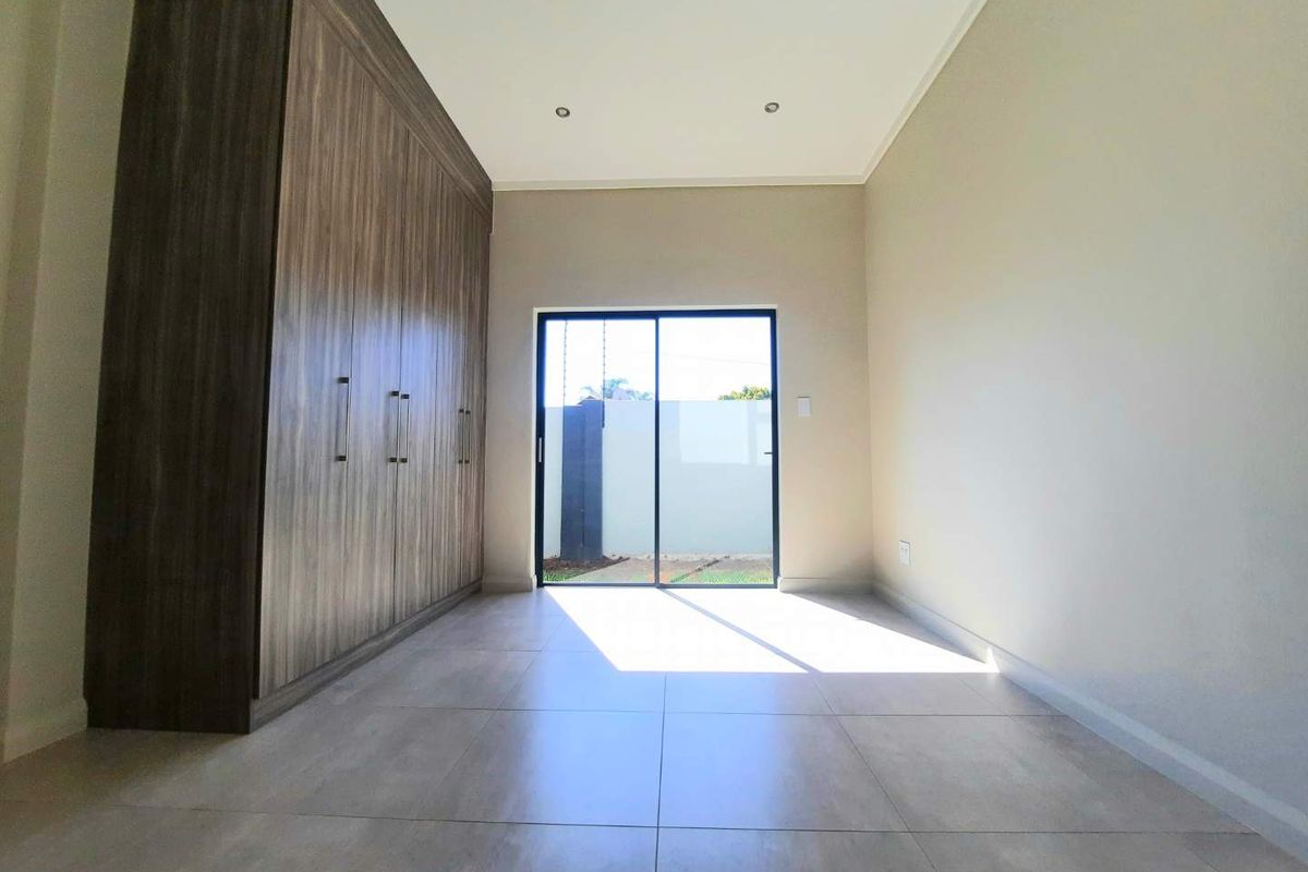 A bright, modern bedroom featuring neutral tiled flooring, extensive built-in wooden cupboards, and a sliding glass door providing access to the exterior.