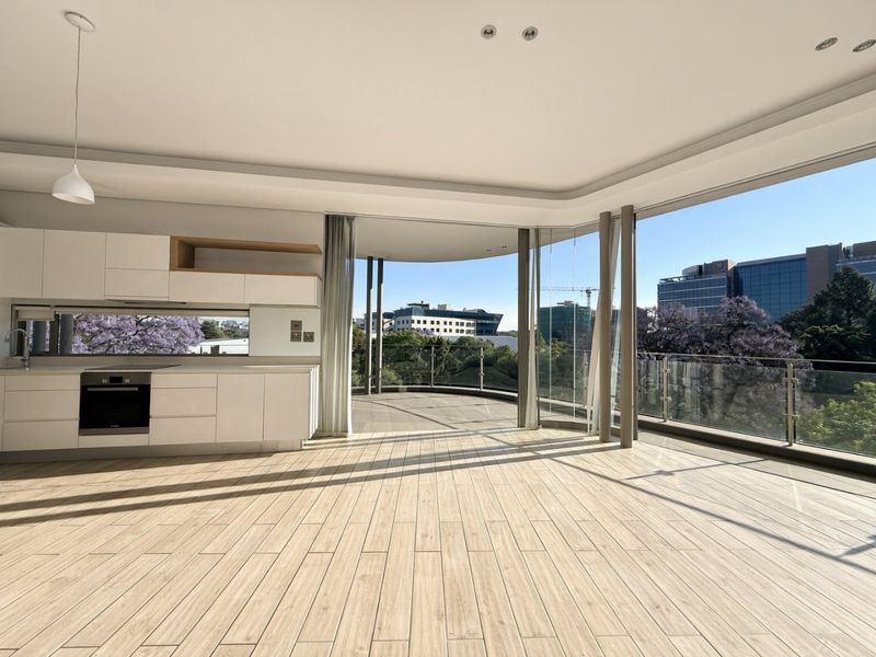 Modern open-plan kitchen and living area featuring sleek cabinetry, integrated appliances, and expansive floor-to-ceiling glass doors opening onto a spacious balcony with city views.