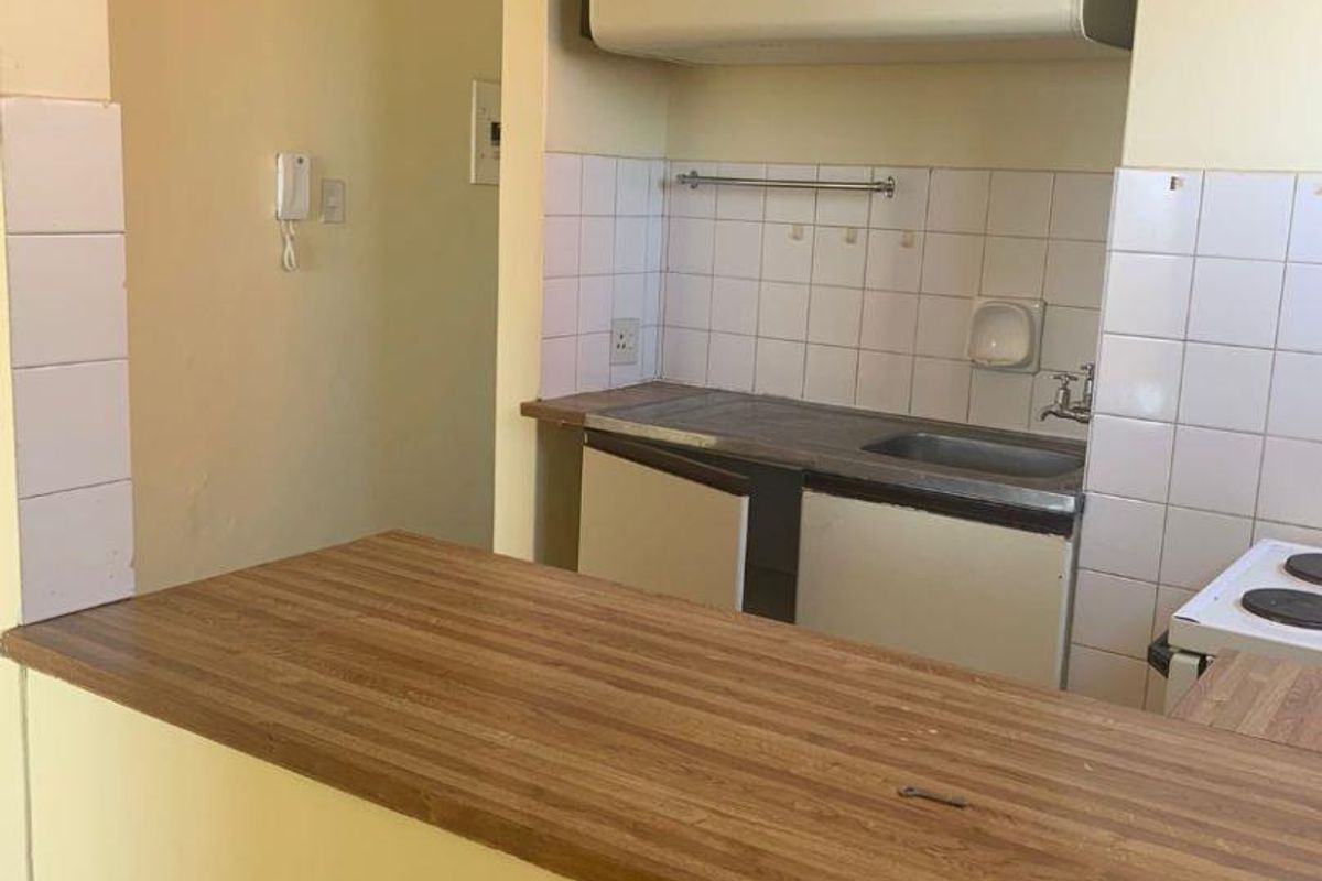 A compact kitchen featuring a breakfast bar, tiled splashback, stainless steel sink, and basic cabinetry, requiring modernization.