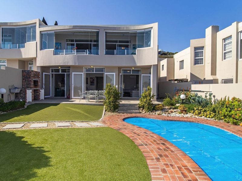 Rear view of the property featuring a swimming pool, artificial lawn, built-in braai area, and an upper-level balcony with glass enclosures.