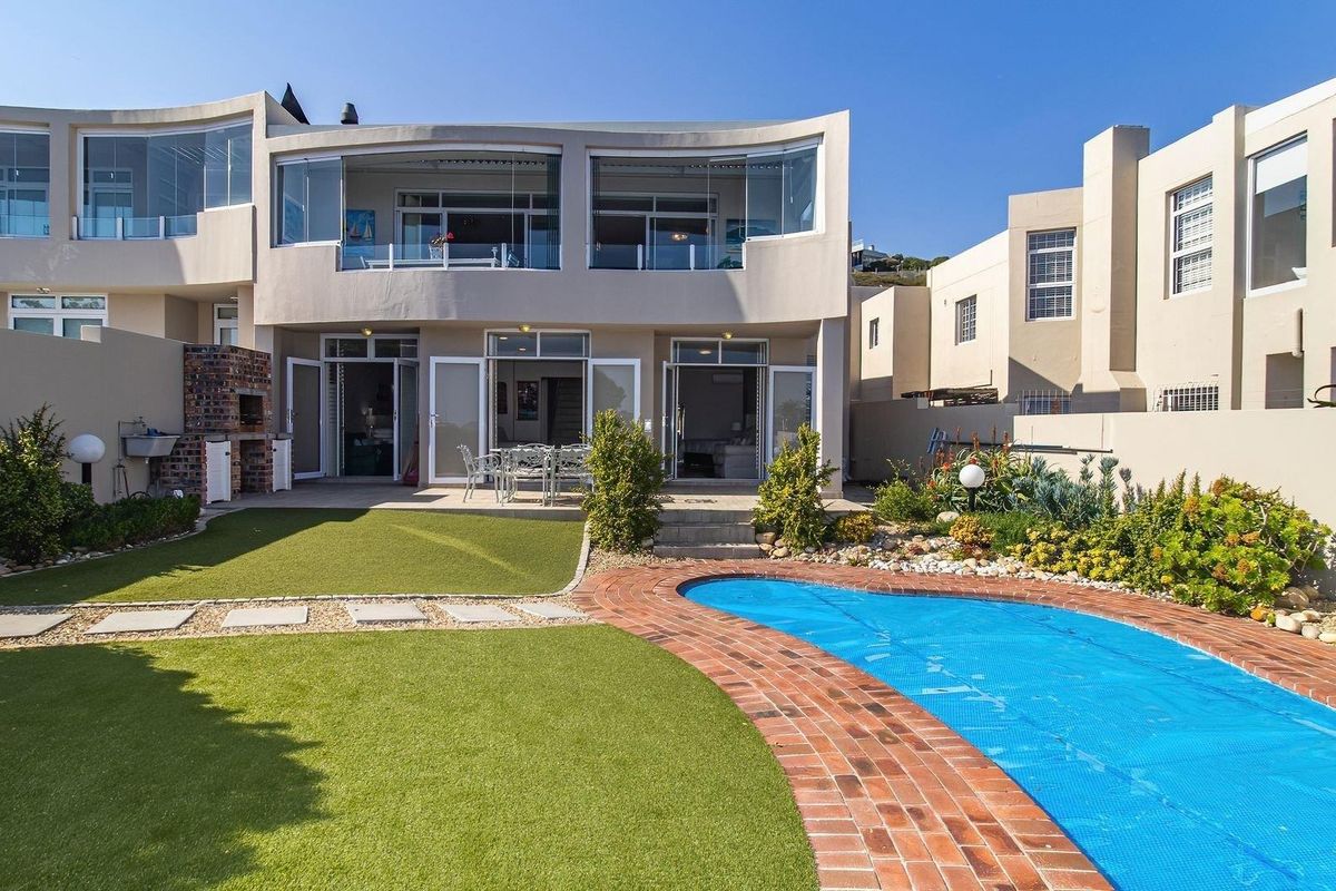 Rear view of the property featuring a swimming pool, artificial lawn, built-in braai area, and an upper-level balcony with glass enclosures.