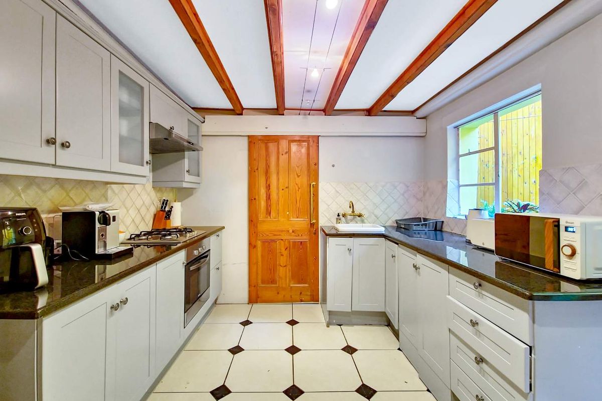 A galley-style kitchen featuring light grey cabinetry, dark stone countertops, a built-in oven, gas hob, and exposed timber ceiling beams.