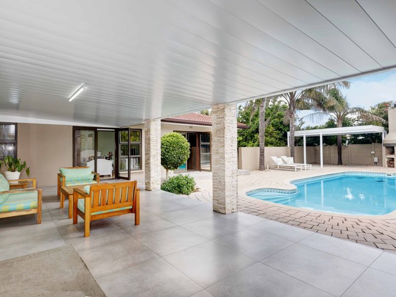 Spacious covered patio featuring stone-clad pillars and tiled flooring, overlooking a well-maintained swimming pool and outdoor entertainment area.