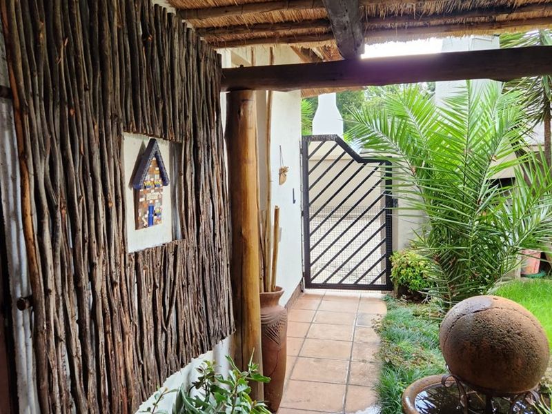 A charming covered patio area featuring a rustic thatched roof, tiled flooring, and a secure metal gate leading to the garden.