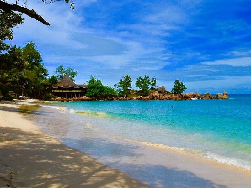 A scenic view of a pristine sandy beach with turquoise waters and a distant coastal structure nestled among tropical trees and rock formations.