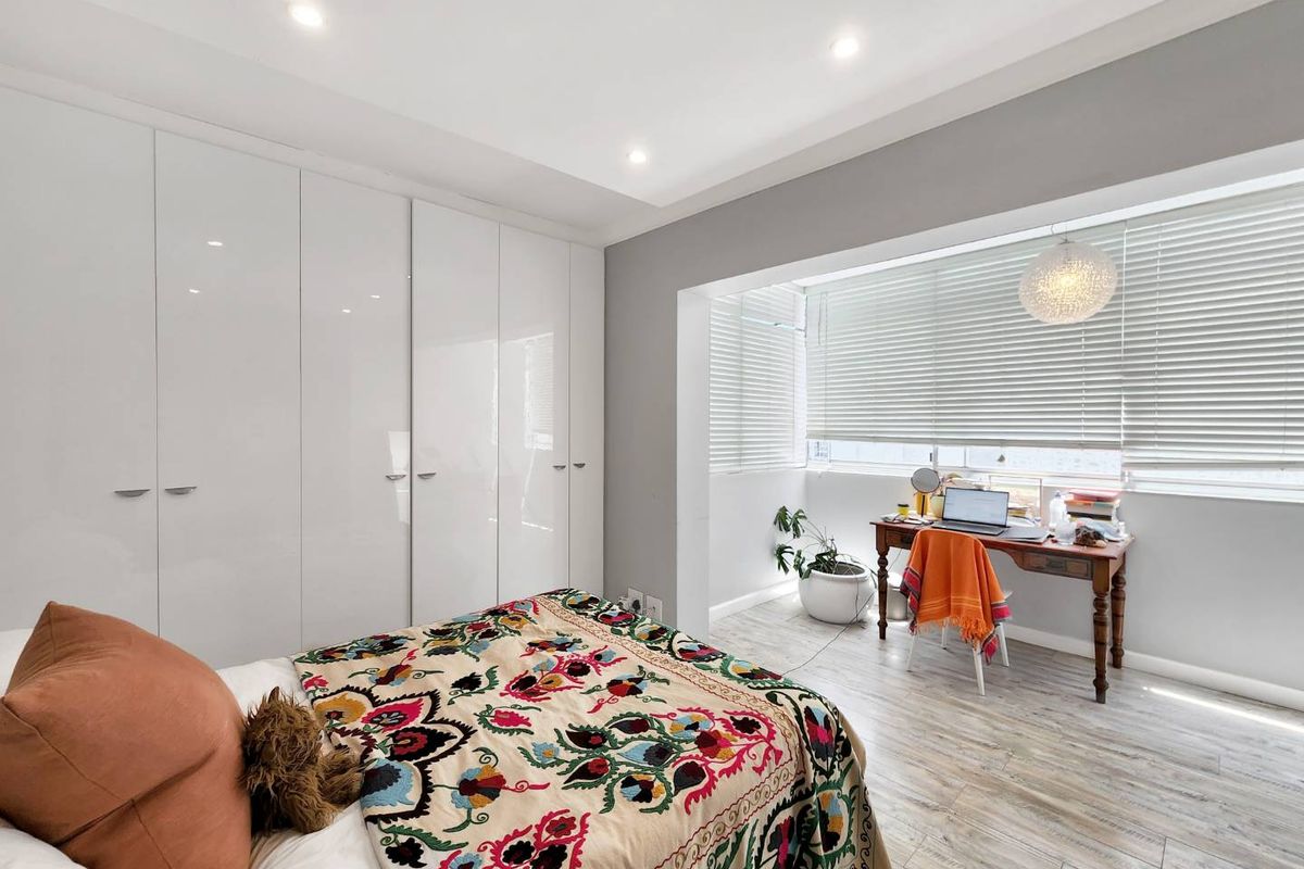 A bright bedroom featuring modern white built-in wardrobes, recessed downlighting, and a dedicated window-side nook with venetian blinds and light-toned laminate flooring.