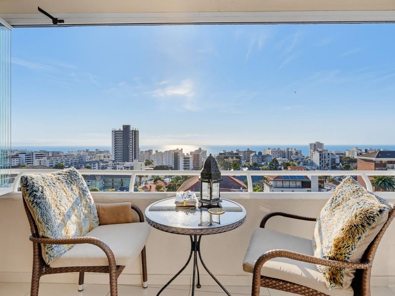 A bright, enclosed balcony featuring glass folding doors and a metal balustrade, offering expansive urban and ocean views.