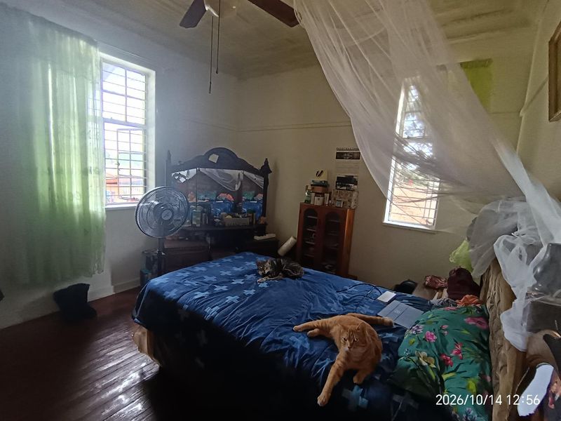 A bedroom featuring timber flooring, a ceiling fan, and security bars on the windows. The room shows signs of wear, including peeling ceiling paint.