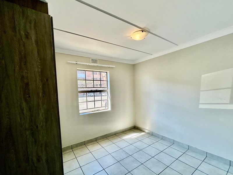 A bright, empty bedroom featuring tiled flooring, a built-in cupboard, and a window with security bars.