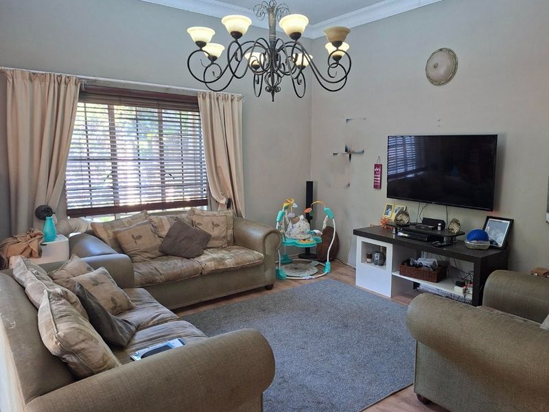 A living room featuring laminate flooring, decorative coving, and a central chandelier, with window blinds providing natural light control.