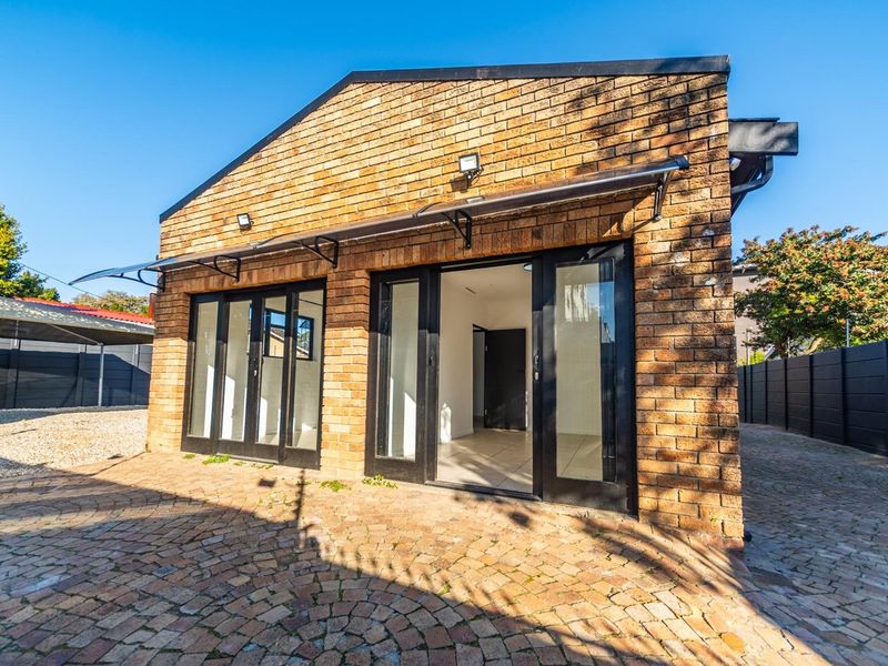A well-maintained face brick exterior featuring black aluminium framed glass doors, modern polycarbonate awnings, and a paved courtyard area.