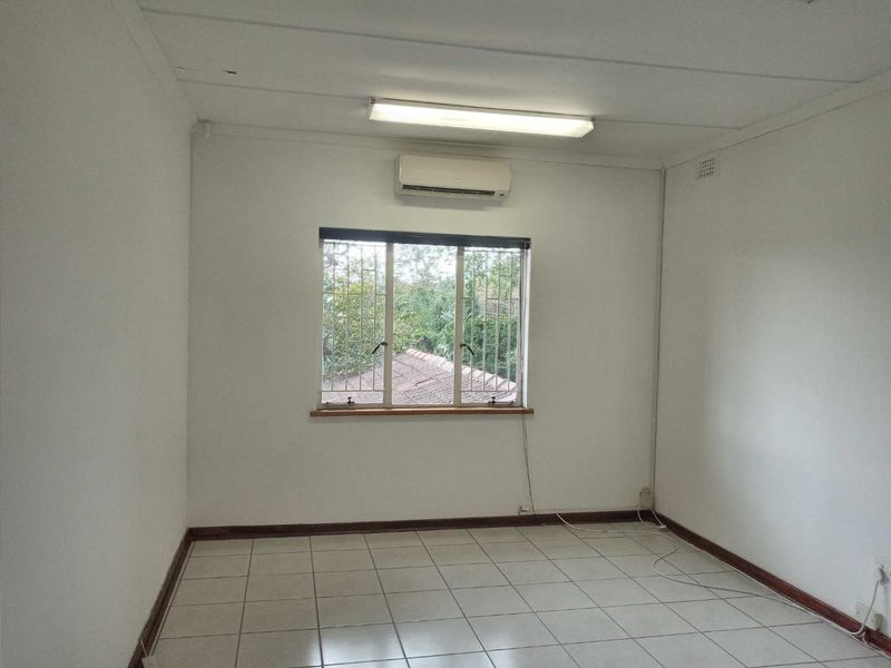 A clean, empty office space featuring tiled flooring, a split-system air conditioner, and a window with security bars.