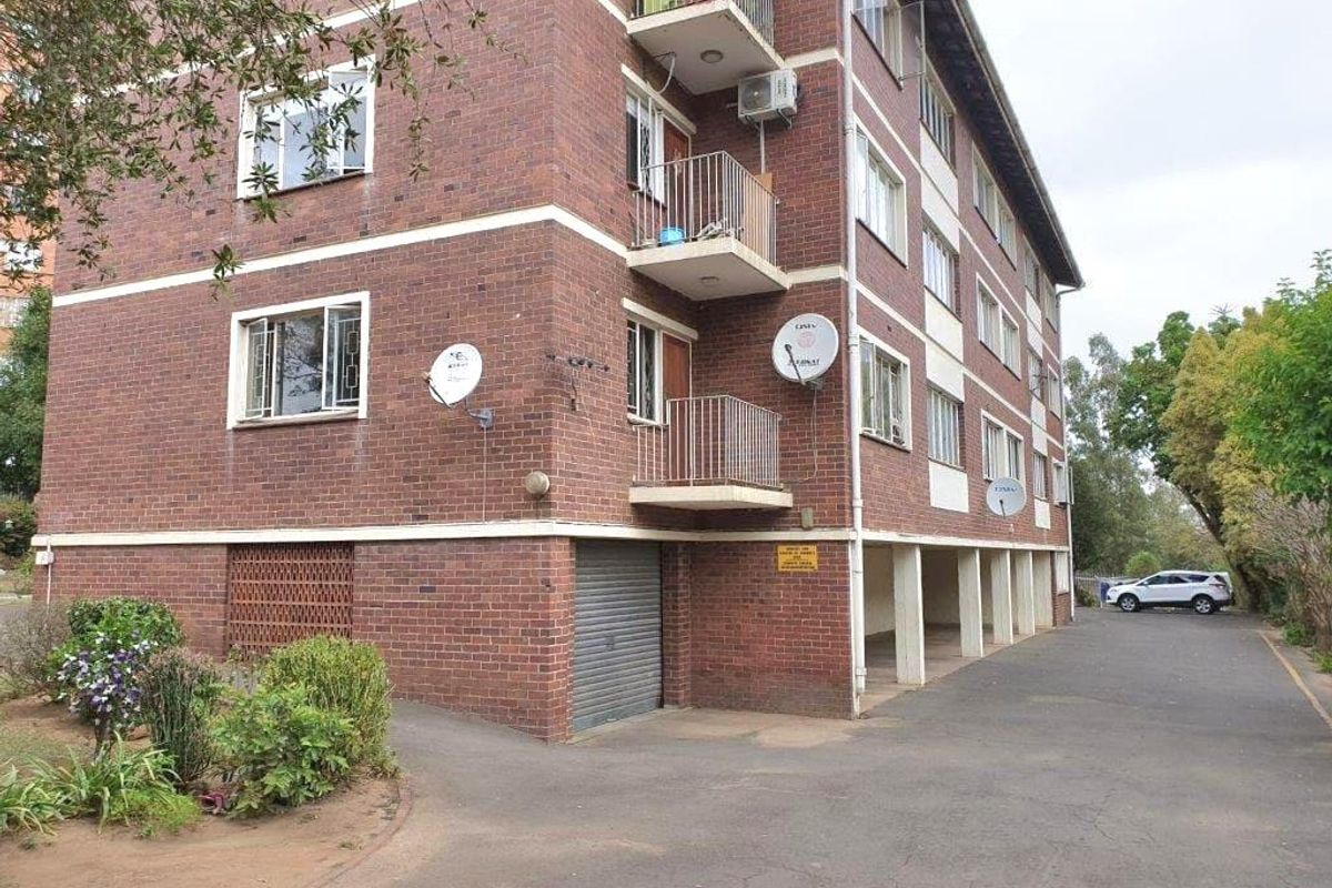 Exterior view of a multi-story brick apartment building featuring private balconies, security bars, and an asphalt driveway with ground-level parking access.
