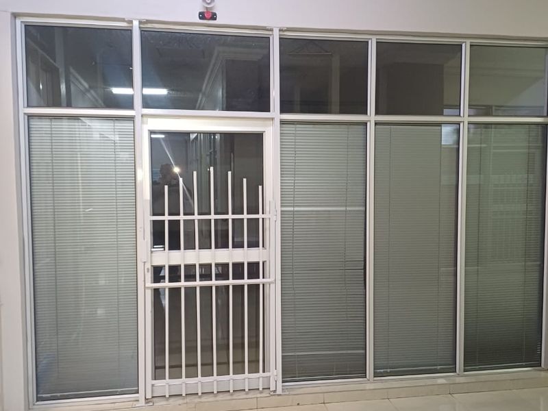 A clean, empty office space featuring large aluminium-framed glass partitions with integrated blinds, a secure entrance gate, and tiled flooring.