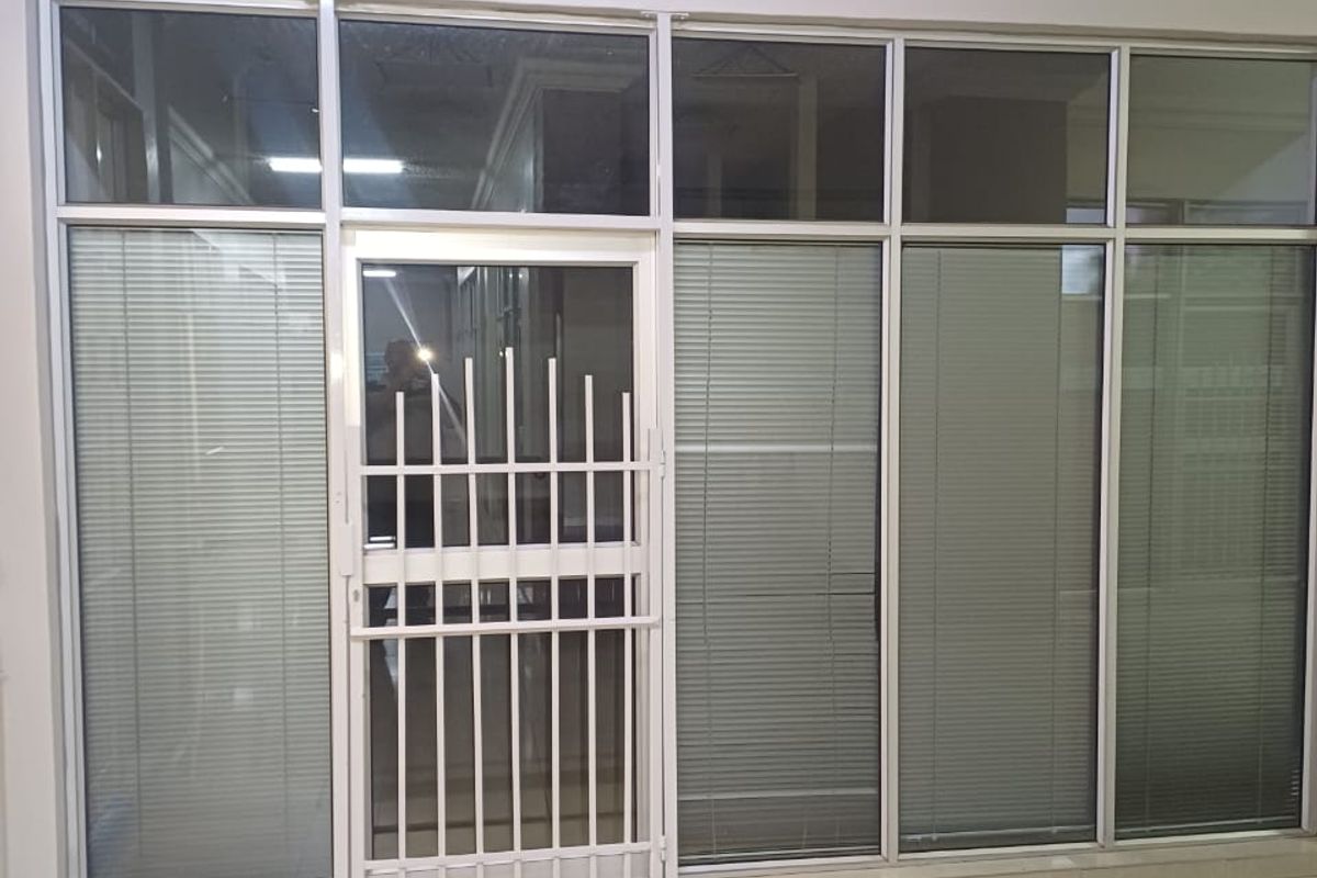 A clean, empty office space featuring large aluminium-framed glass partitions with integrated blinds, a secure entrance gate, and tiled flooring.