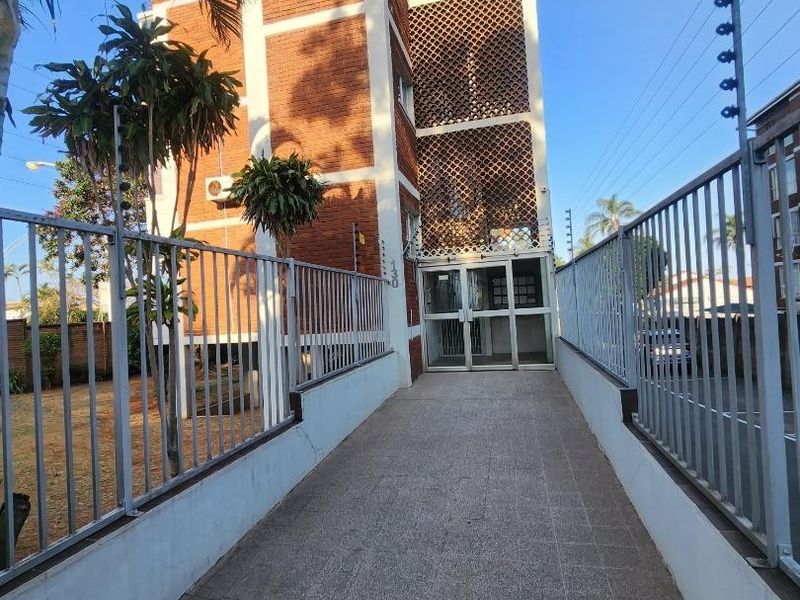 Front entrance view of the apartment building featuring a paved walkway, secure metal fencing, and a distinctive brick and breeze block facade.