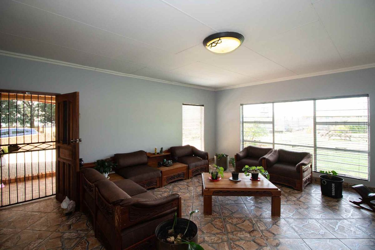 A spacious living room featuring tiled flooring, large windows with blinds, and a security gate at the entrance.