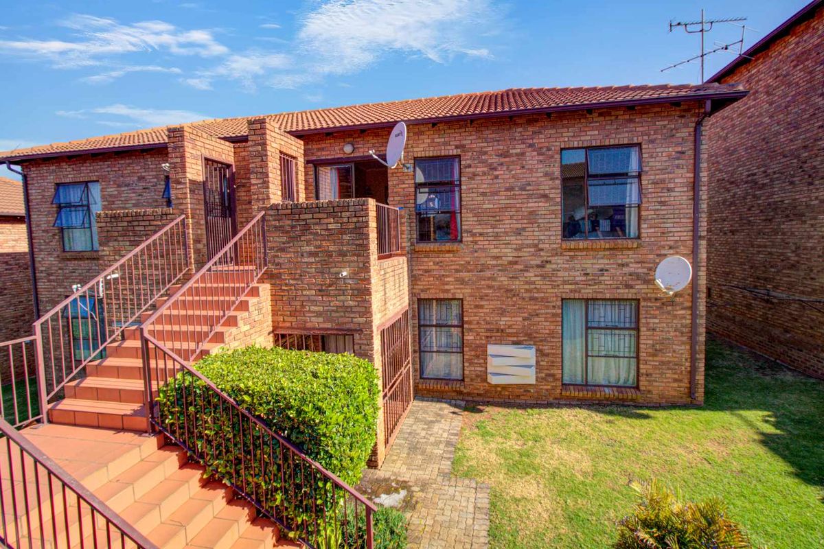Exterior view of a two-story brick apartment building featuring an external staircase, tiled balcony, and surrounding lawn area.